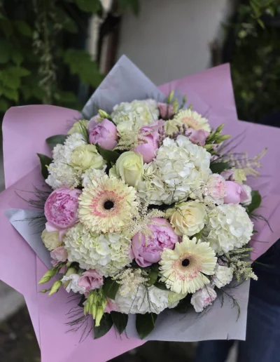 Un bouquet de fleurs composé de pivoines roses, d'hortensias blancs, de roses et de gerberas pâles au centre rosé, le tout enveloppé dans du papier rose