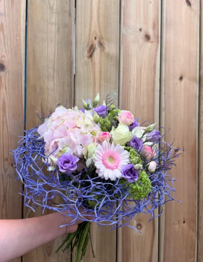 Un bouquet rond et harmonieux qui associe des hortensias rose pâle, un gerbera rose, des lisianthus violets et des roses rose tendre et crème, le tout reposant sur une structure fine de branches bleues
