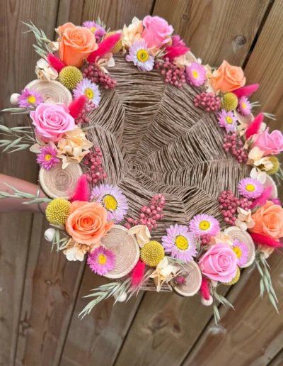 Bouquet stabilisé monté sur une structure tressé, rondin de bois, composé de roses orange , de fleurs jaunes, de petites marguerites, de fleurs fuchsia et de touches de verdure