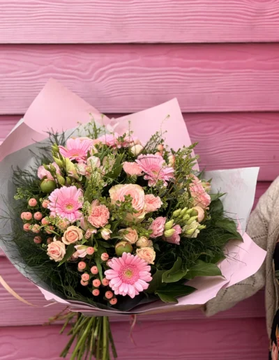 Composition florale vibrante associant des germini roses bonbon, de petite roses peach, des oeillets, et des baies d'hypericum qui contrastent avec un feuillage verdoyant, le tout enveloppé dans du papier rose